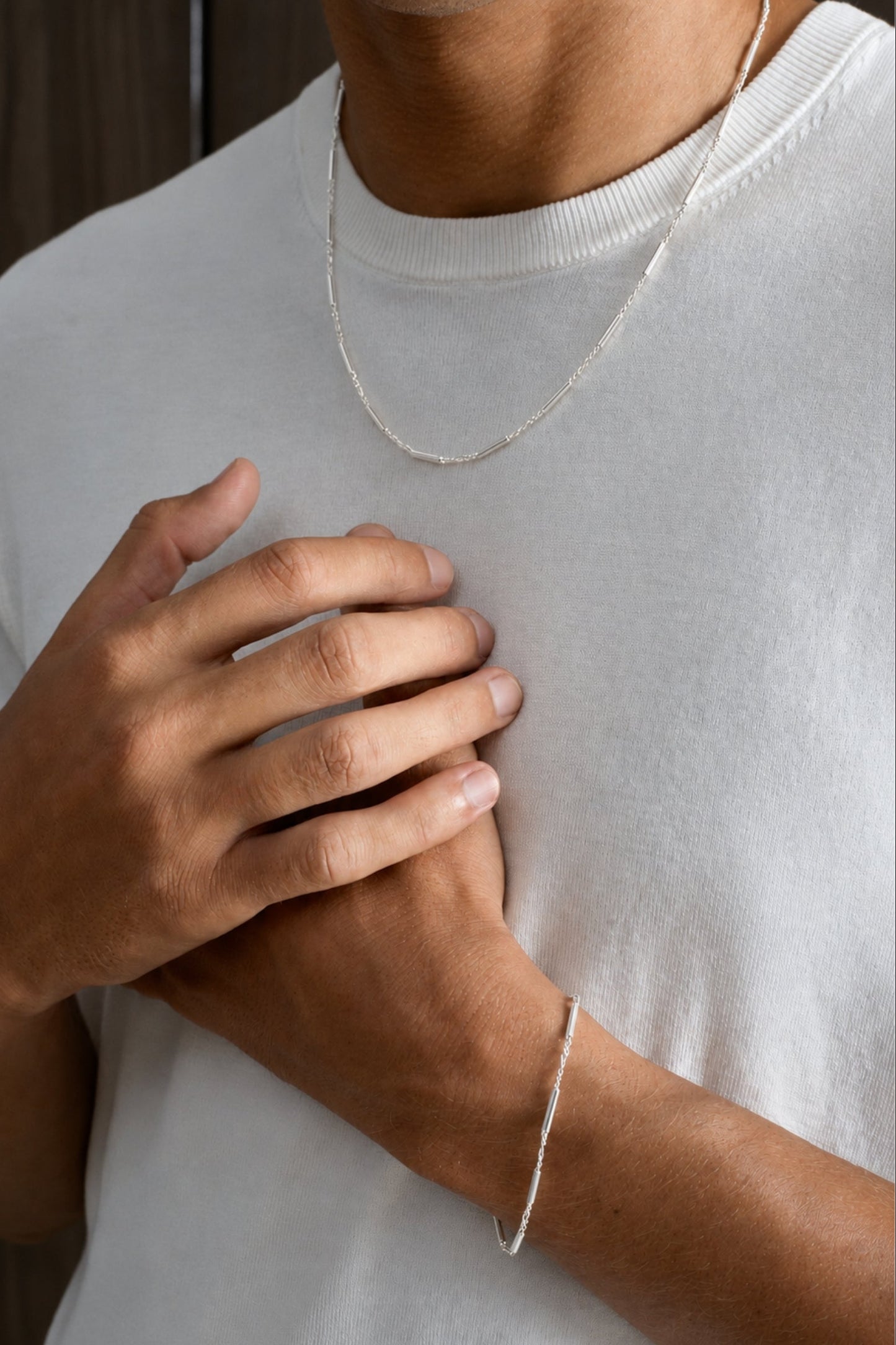 A close-up of a person wearing a delicate silver chain necklace and a matching thin silver bracelet, paired with a white crew-neck t-shirt.