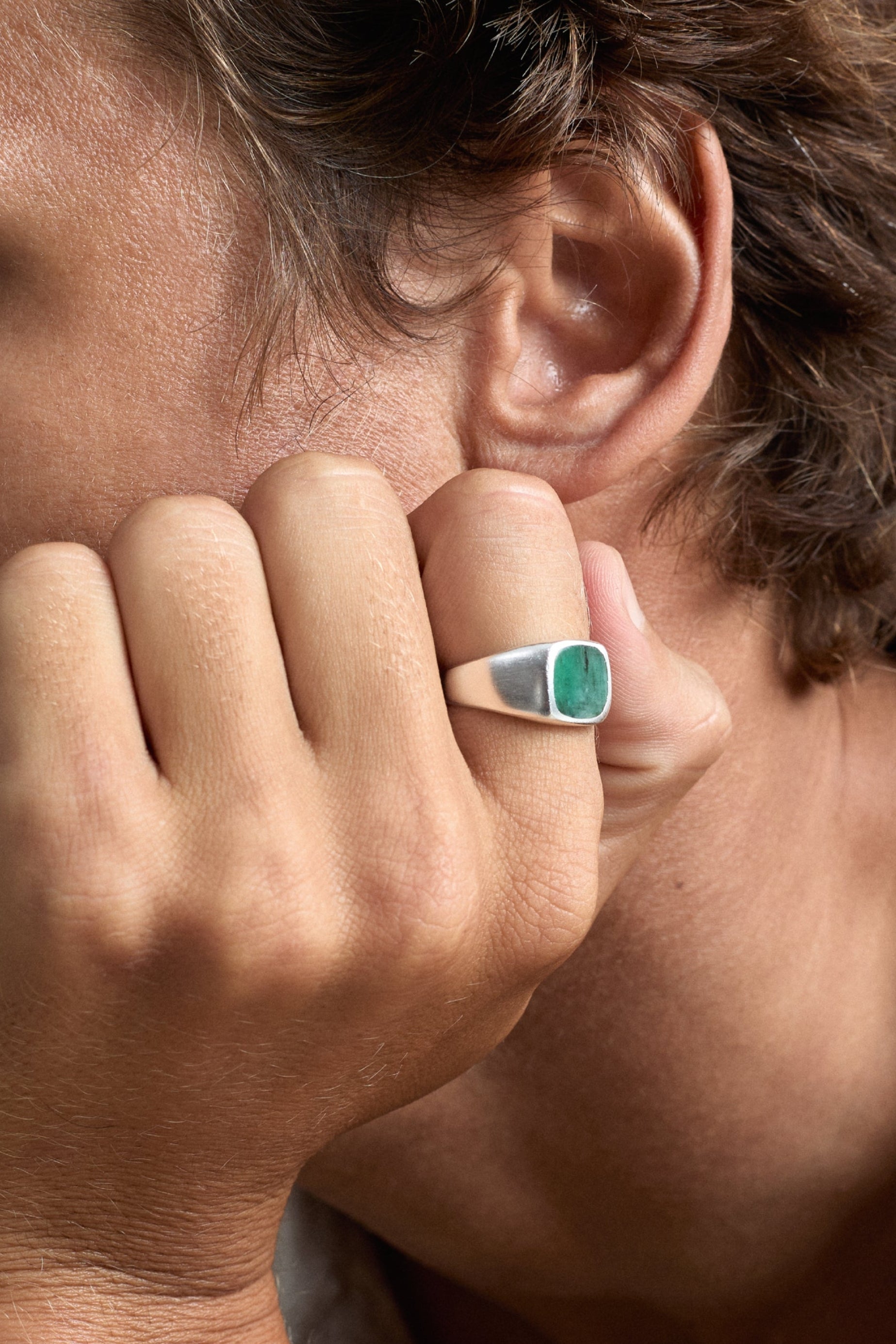 Close-up of a person wearing a ring with a green gemstone, holding their hand to their face.