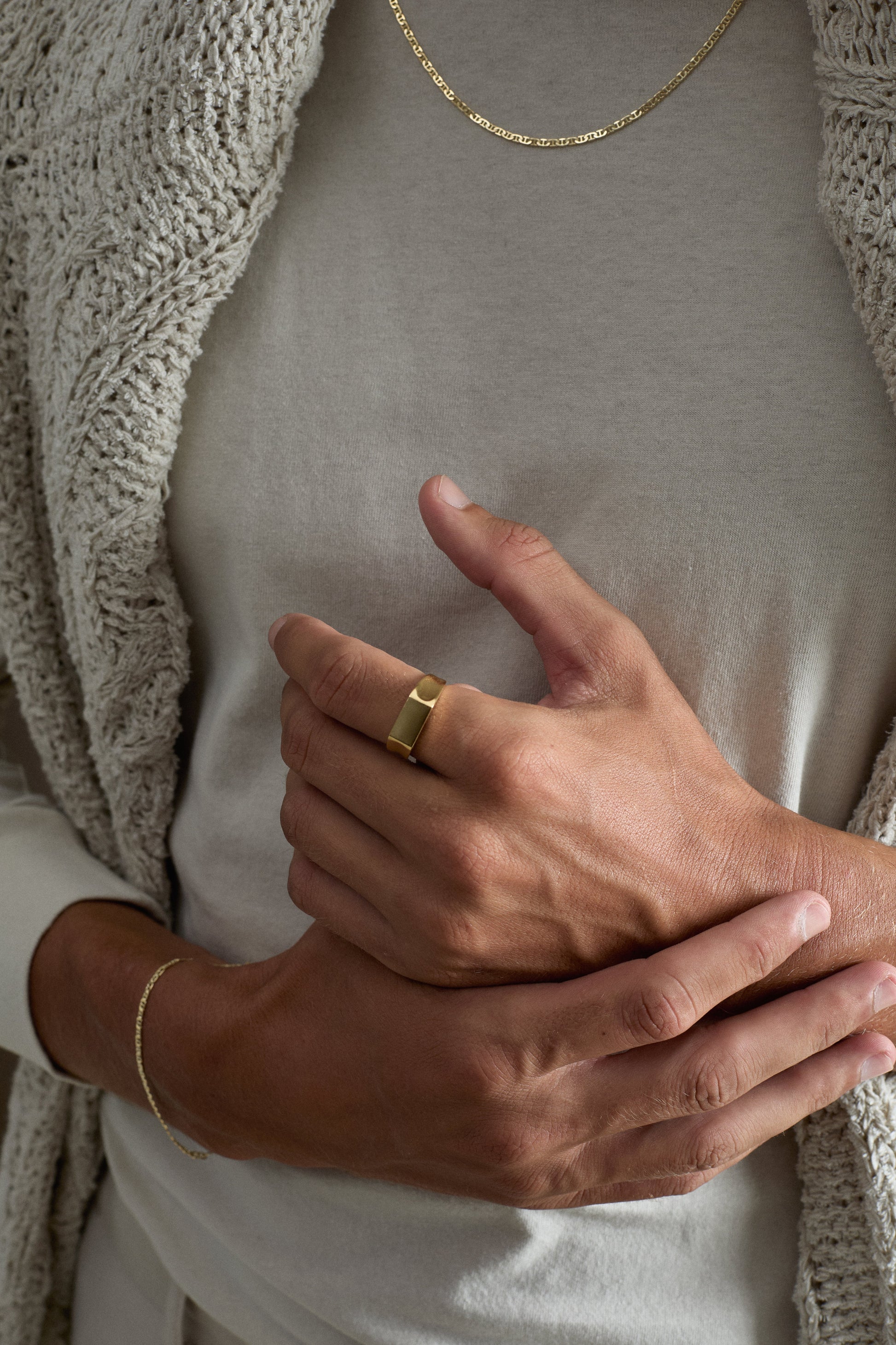 Close-up of a person wearing a gold ring and gold necklace, with a neutral background.
